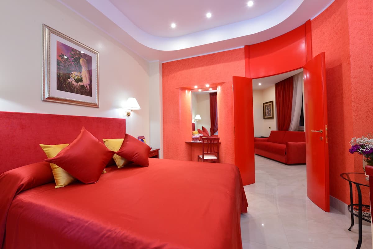 A hotel room with a red bed, a desk with a chair, a red sofa, and a mirror.