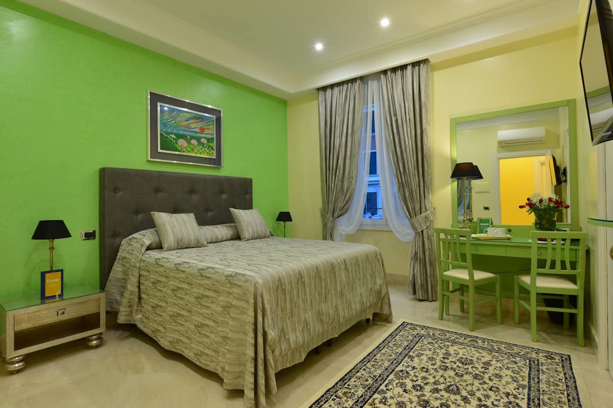 A bedroom with a bed, a desk, chairs, a lamp, a picture on the wall, and a window with curtains.