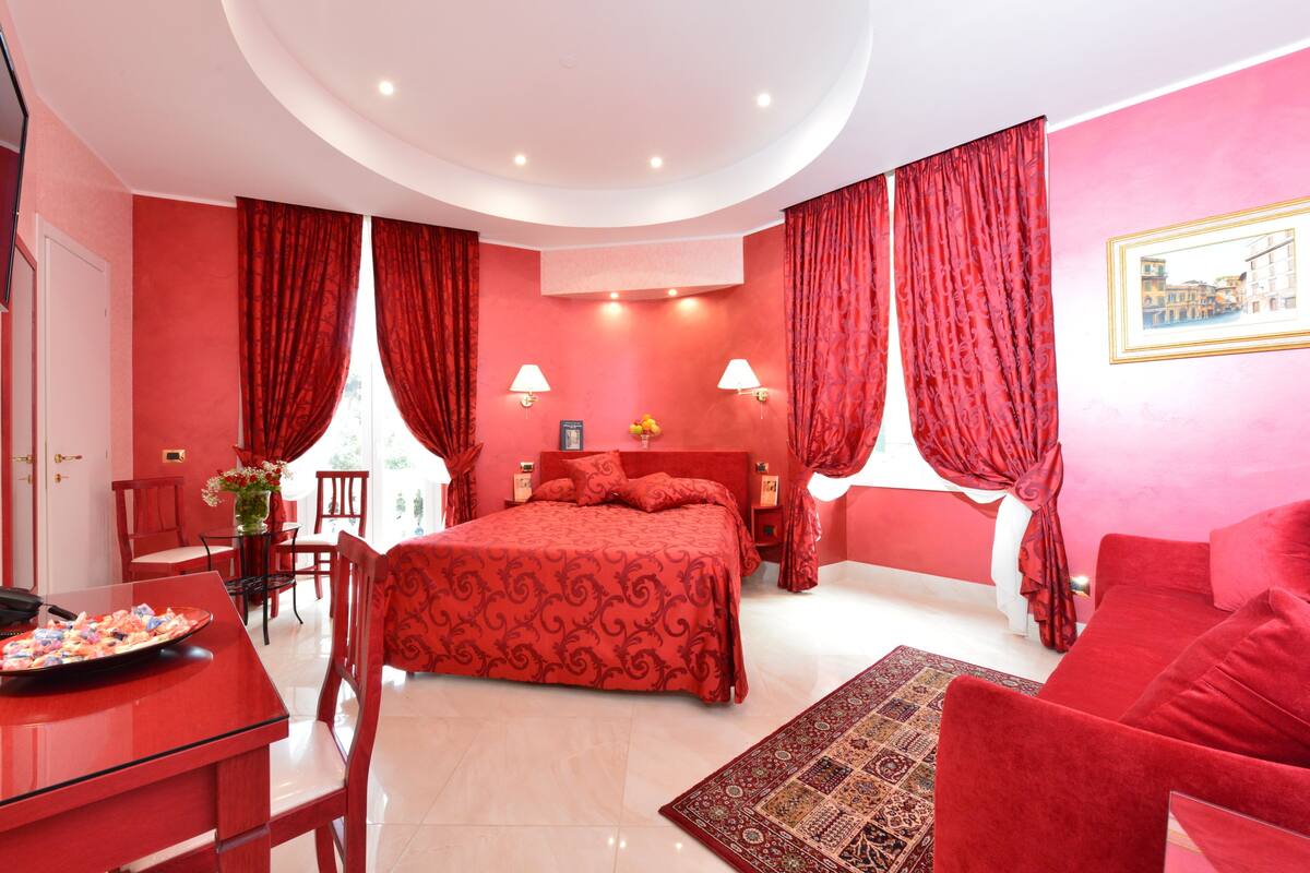 A red-themed bedroom with a bed, two bedside tables, a desk with a chair, and a red sofa.