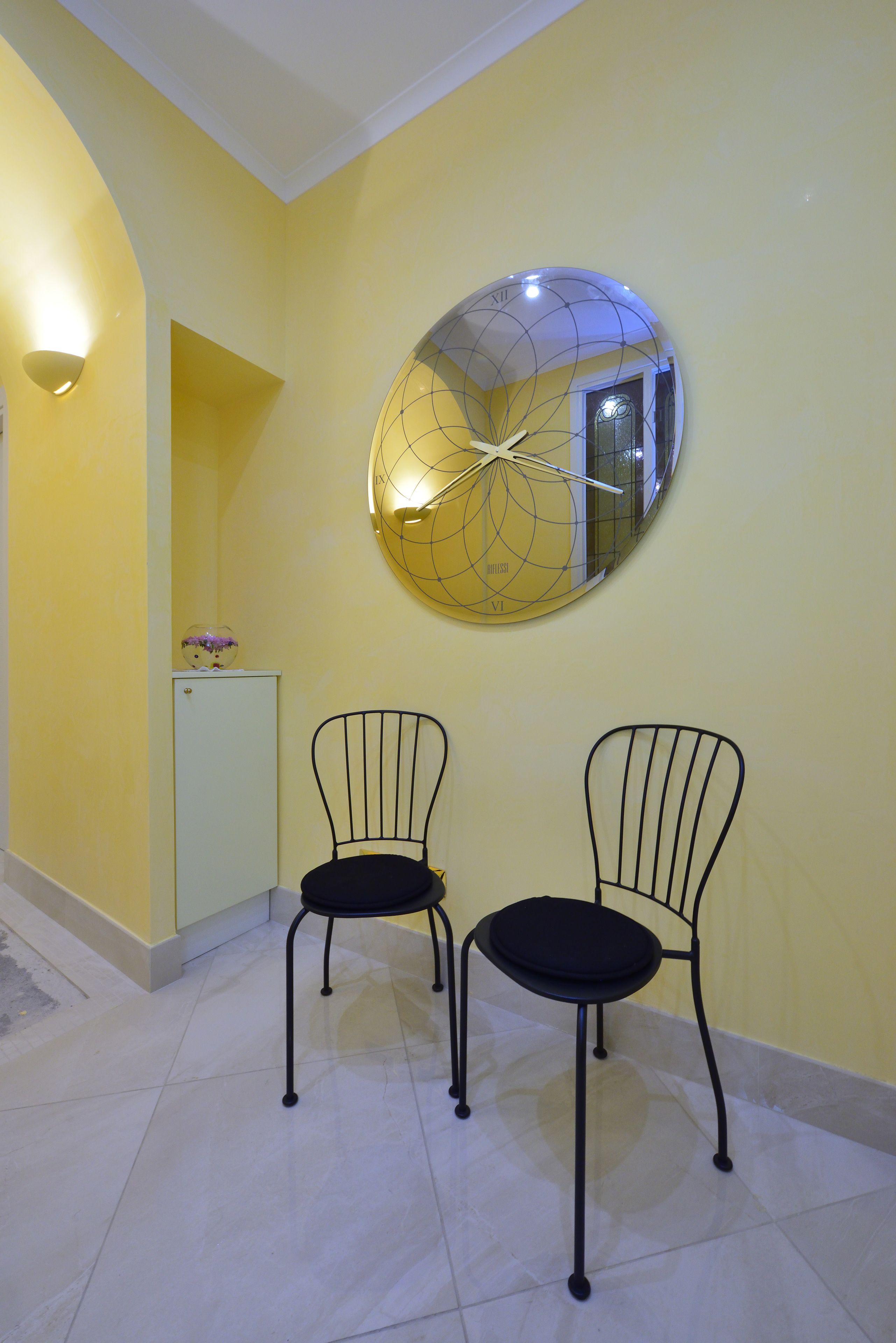 Una habitación con paredes amarillas, un espejo redondo, dos sillas negras y un pequeño armario blanco.