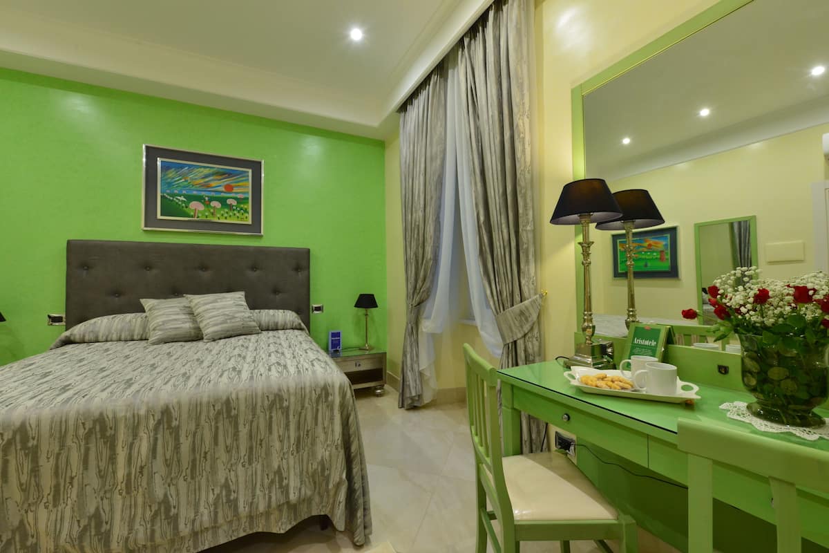 A hotel room with a bed, a desk with a chair, a mirror, and a vase with flowers.