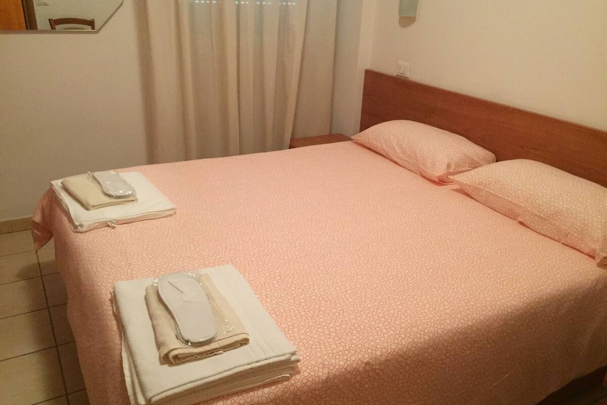 Una cama bien hecha con una colcha estampada rosa y almohadas, un cabecero de madera, y una mesita con toallas dobladas y un teléfono.