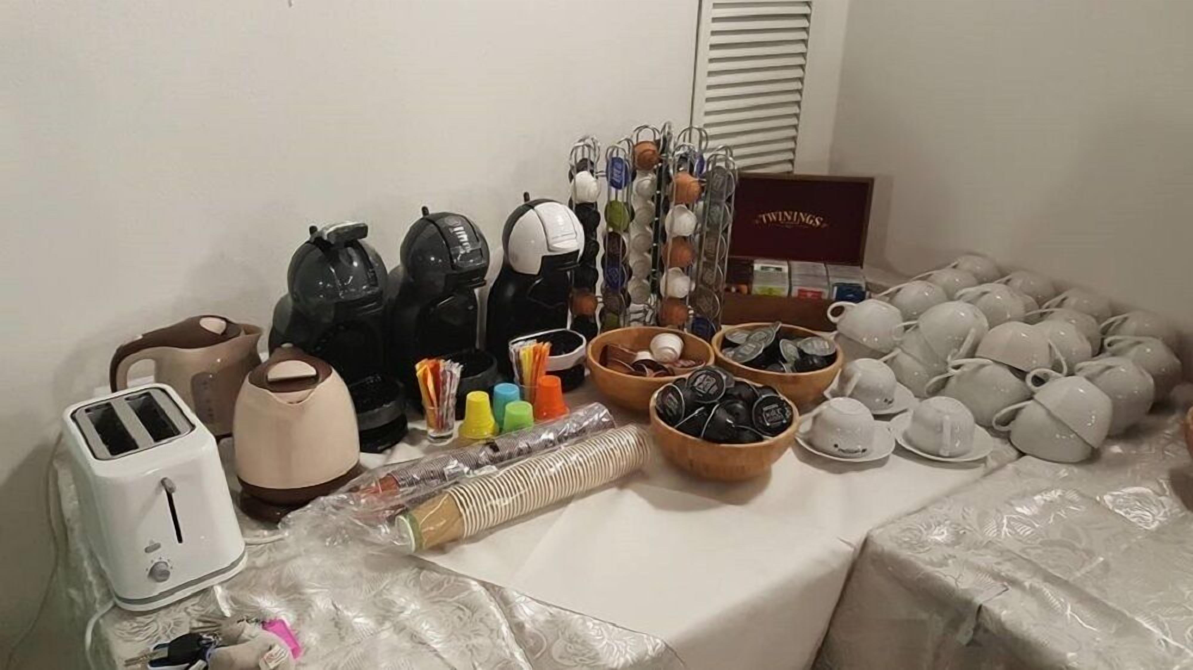 Ein Zimmer mit einem Tisch, auf dem verschiedene Gegenstände liegen, darunter ein Toaster, Wasserkocher, Helme und eine Schachtel mit der Aufschrift 'TWININGS'.