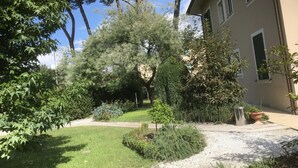 Un jardín bien cuidado con diversas plantas y árboles, un sendero de grava que conduce a un edificio de fachada color crema y ventanas con contraventanas.