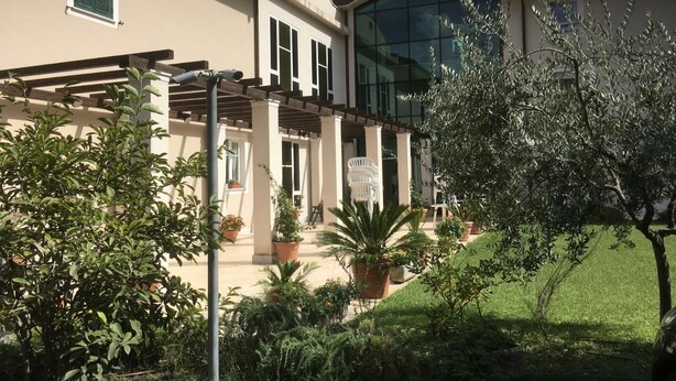 Una casa moderna con fachada de vidrio curvo, un porche cubierto con mobiliario exterior y un jardín bien cuidado.