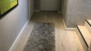 Käytävän lattialla on harmaa, paksukuitinen matto, sen päässä on valkoinen ovi ja seinällä on kehystetty taulu.
