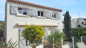 A two-story building with white walls and a tiled roof. There are multiple windows with shutters, a small garden area with various plants, and a fence surrounding the property.
