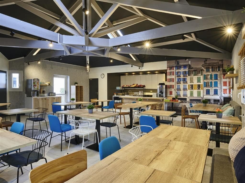 Zona pranzo moderna con tavoli in legno e sedie blu, cucina sullo sfondo e un originale design del soffitto.