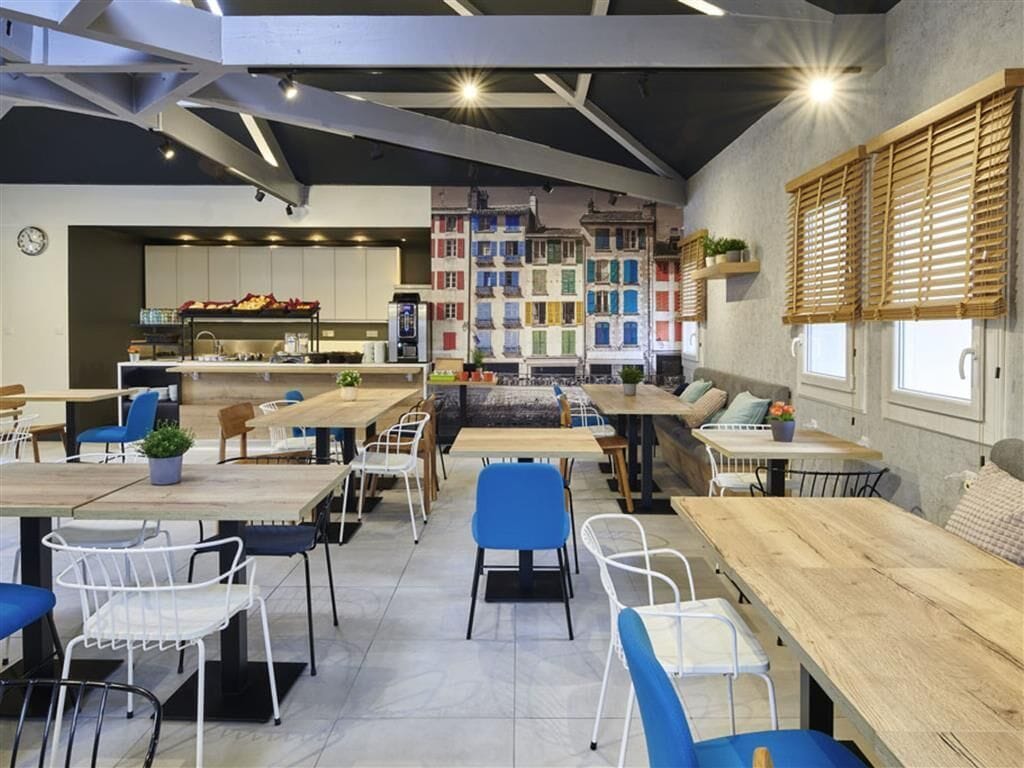 A modern cafe with wooden tables and chairs, a kitchen area, and colorful buildings in the background.