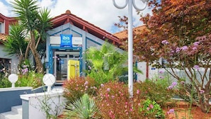Un bâtiment à la façade bleue et blanche, une enseigne indiquant « ibis budget », et un jardin bien entretenu avec diverses plantes et fleurs.