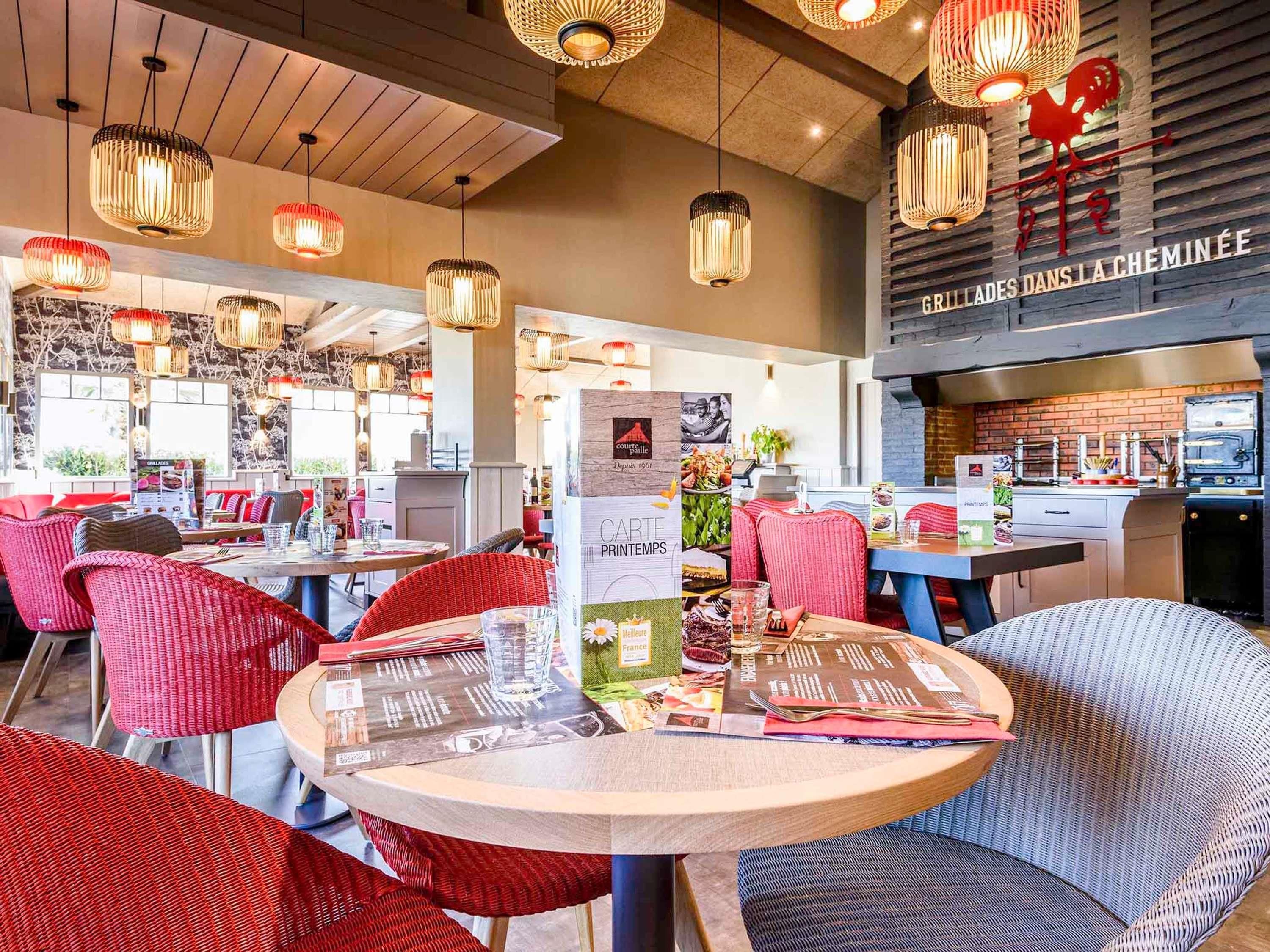 A restaurant interior with round tables, red and grey upholstered chairs, hanging wicker lamps, and a visible kitchen area with a brick wall.