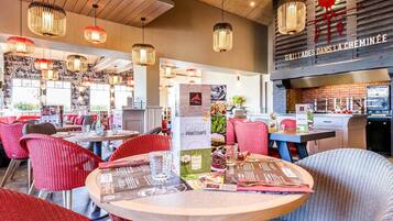 A restaurant interior with round tables, red and grey upholstered chairs, hanging wicker lamps, and a visible kitchen area with a brick wall.
