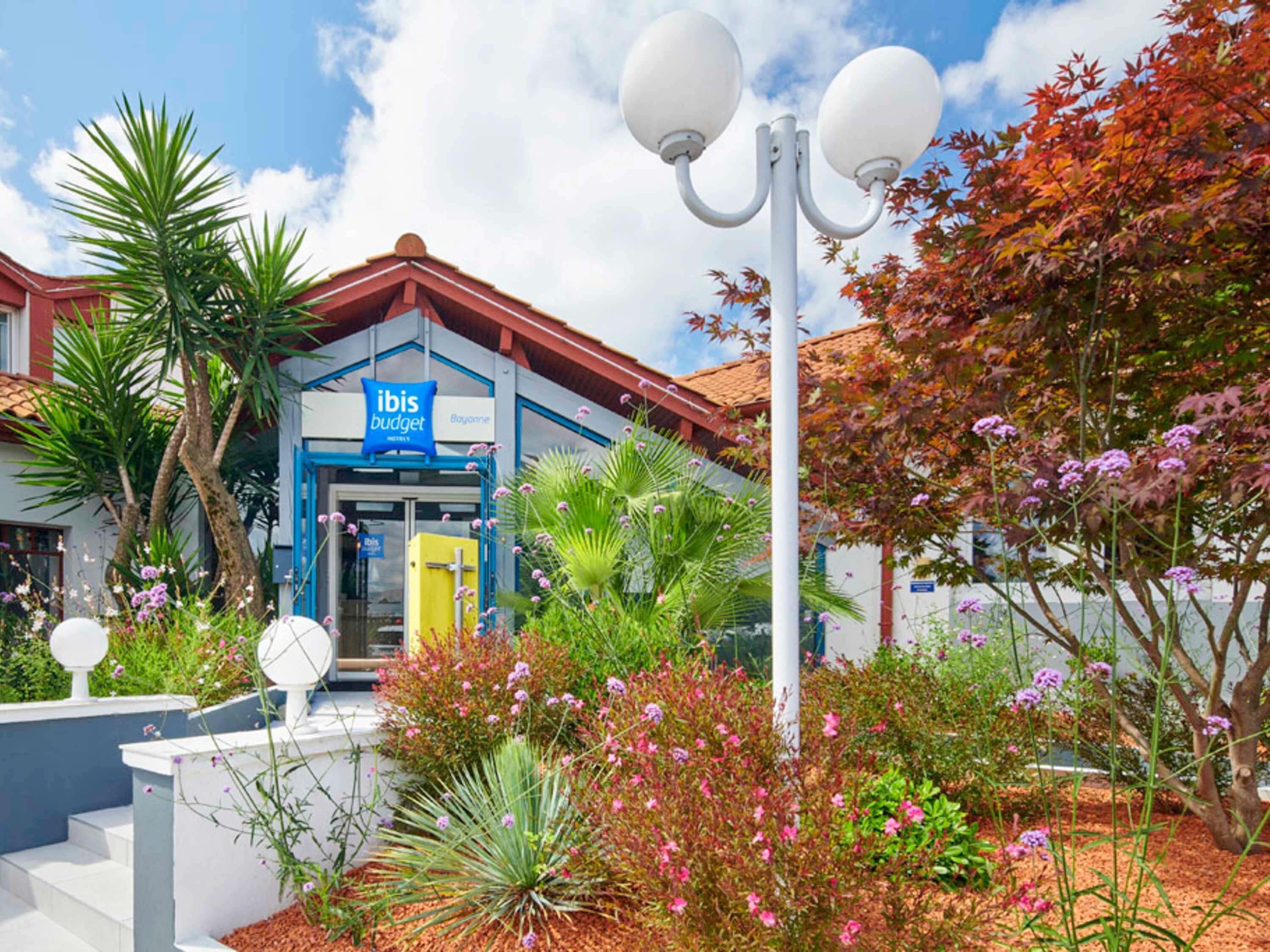 A hotel with a blue and white facade, a visible entrance, and a landscaped garden with various plants and flowers.