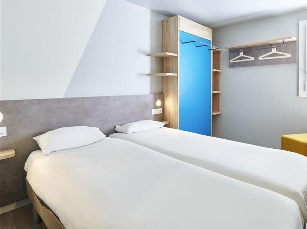 A modern hotel room with two beds, a built-in wardrobe with shelves, and a wall-mounted coat rack.