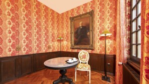 Una stanza con carta da parati decorata, un tavolo tondo con vassoio, una sedia, una lampada da terra e un grande quadro incorniciato appeso al muro.