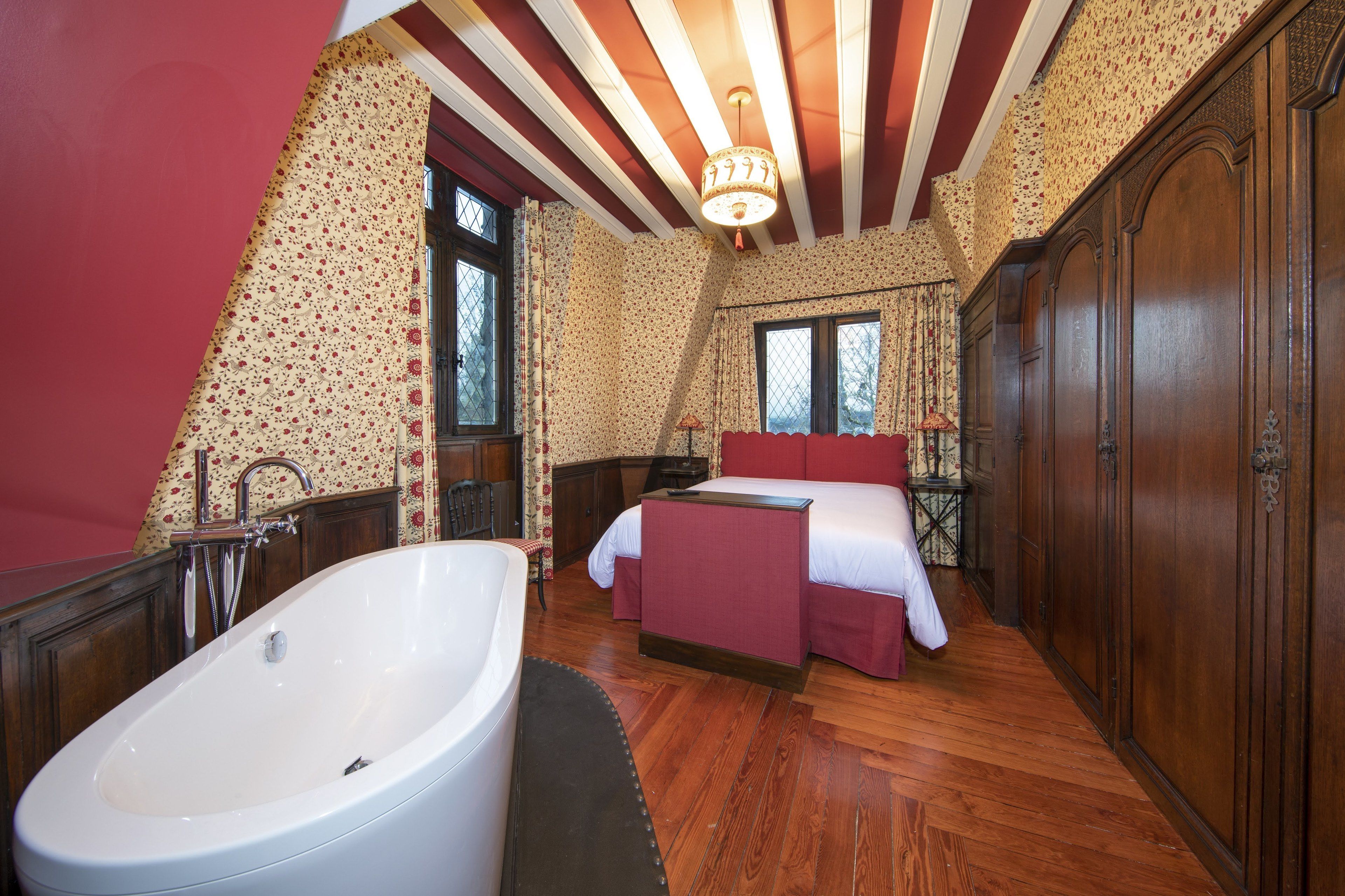 A bedroom with a large white bathtub, a bed with red bedding, and a wooden wardrobe.