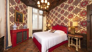 A bedroom with a large bed, a red headboard, a chandelier, patterned wallpaper, and a portrait on the wall.