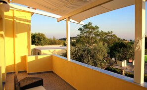 A balcony with a view of trees and buildings, a wicker chair, and a tiled floor. - Belvedere (Procida)