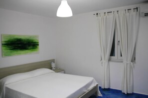 A bedroom with a bed, a nightstand, a wall-mounted lamp, and a window with curtains. - Belvedere (Procida)