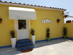 A yellow building with a white awning labeled "Belvedere PROOF." There are steps leading to the entrance, potted plants, and a wall-mounted decorative piece. - Belvedere (Procida)