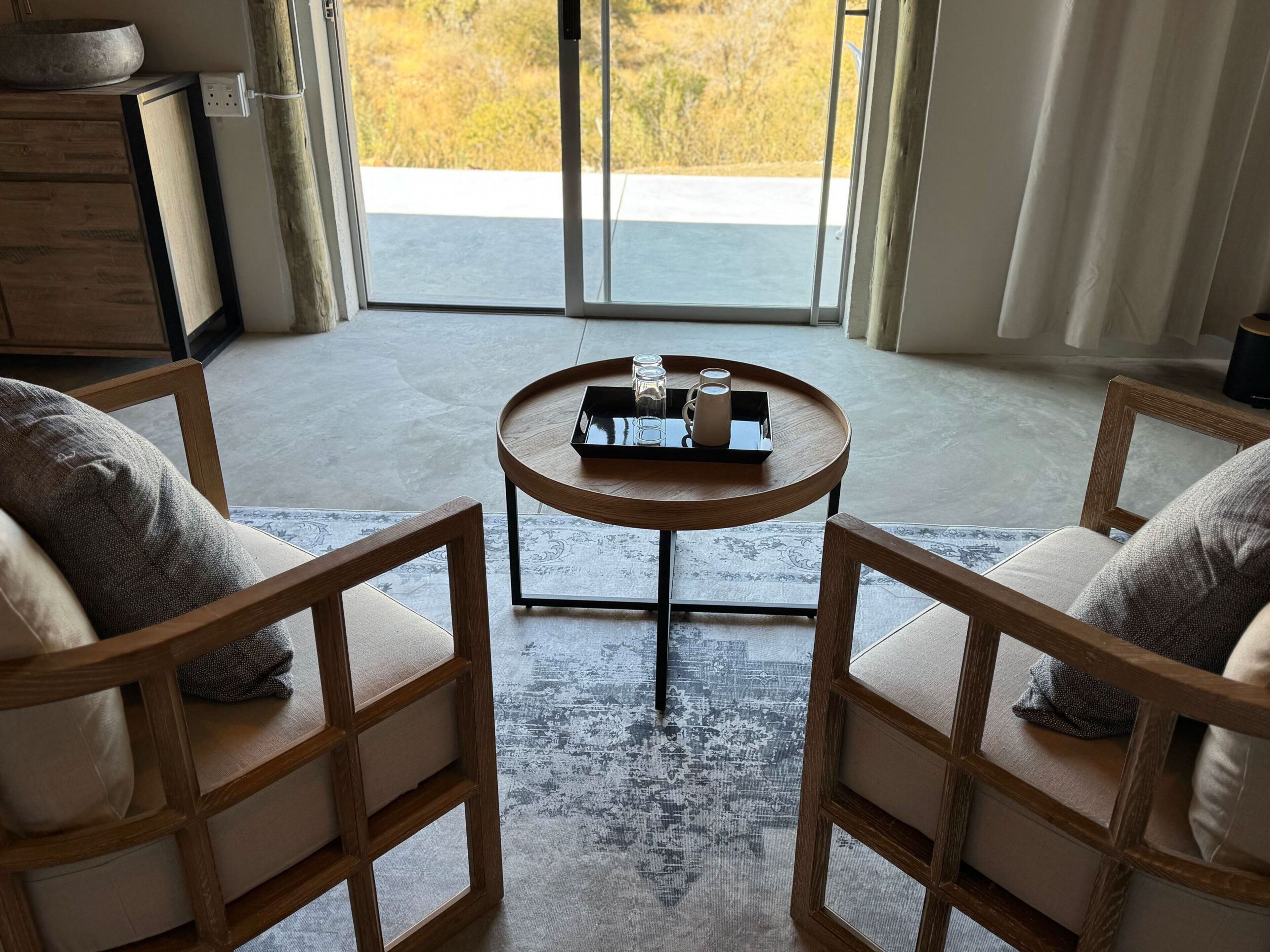 A room with two wooden chairs, a small round table with a tray, and a view of a road and greenery outside.