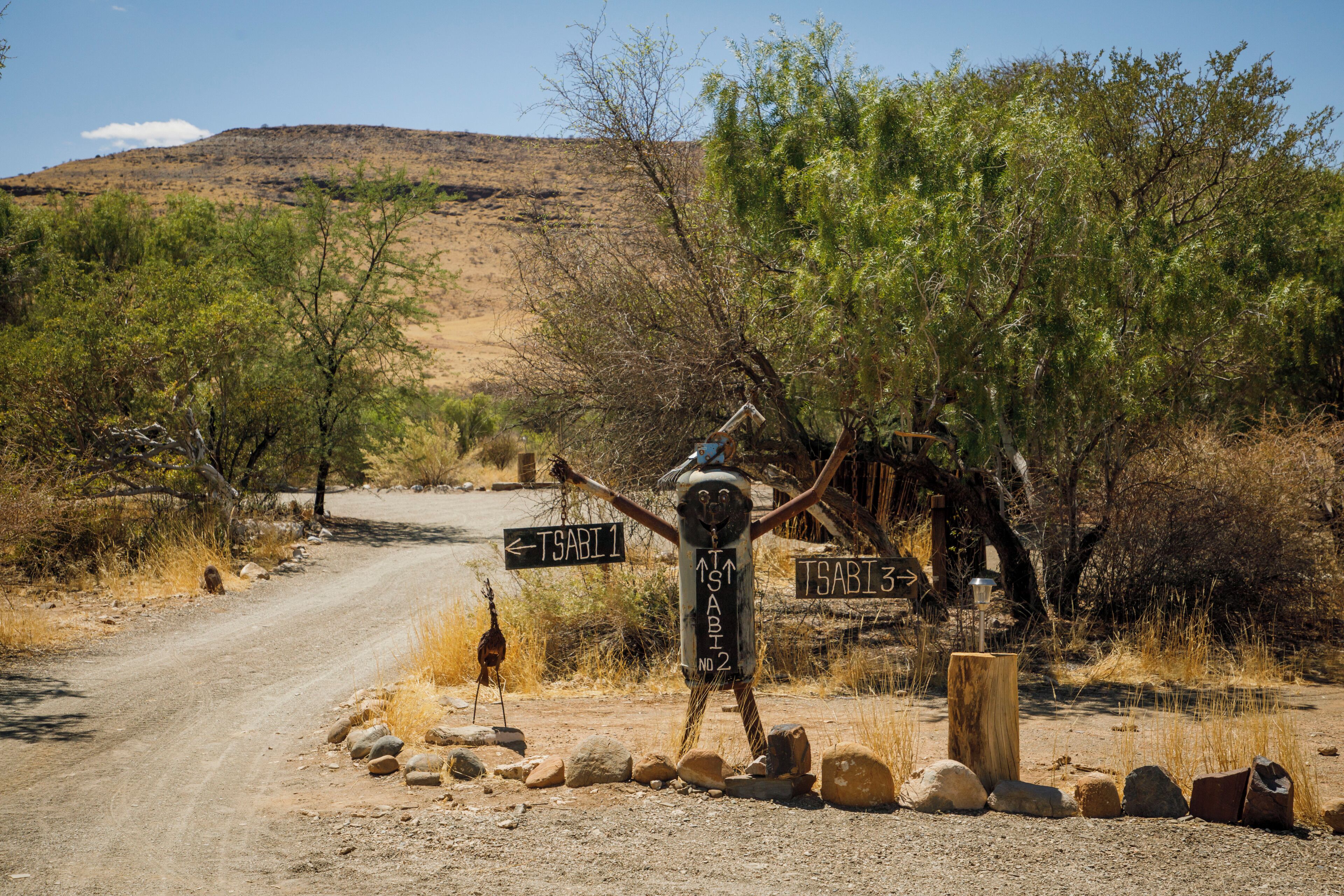 A rustic signpost with directional arrows and a wooden post, surrounded by dry vegetation and a dirt road.