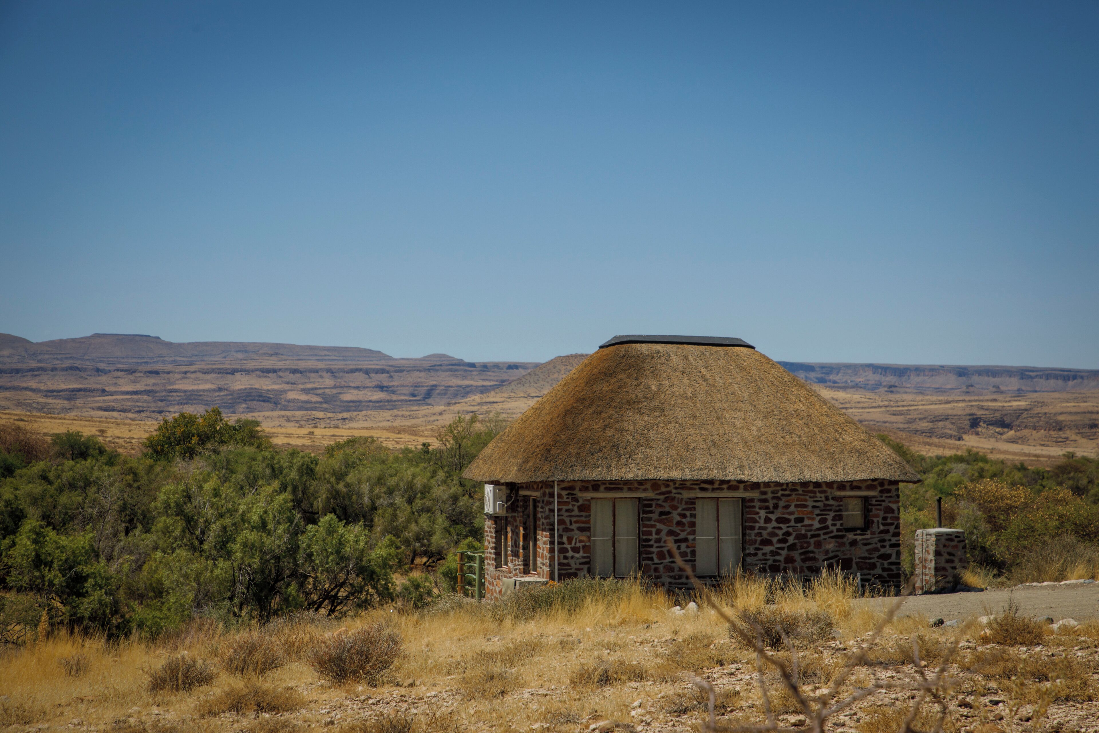 Ein kleines Steinhaus mit einem Reetdach, umgeben von trockenem Gras und vereinzelten Bäumen, vor einer Kulisse ferner Hügel.