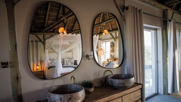 A bathroom with two oval mirrors, a wooden vanity, and a large window with curtains. - Tsauchab River Lodge (Sesriem)