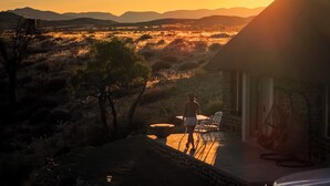 A person walking towards a building with a patio, surrounded by a desert landscape with mountains in the background. - Tsauchab River Lodge (Sesriem)