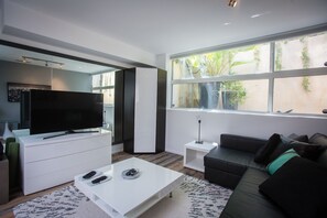 A modern living room with a flat-screen TV, white furniture, a black leather sofa, and large windows offering a view of greenery.