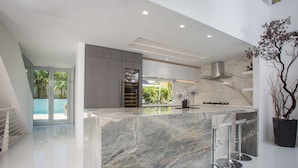 A modern kitchen with a marble island, stainless steel bar stools, and a view of the outdoors. - Modern Del Mar Beach Home (Del Mar)