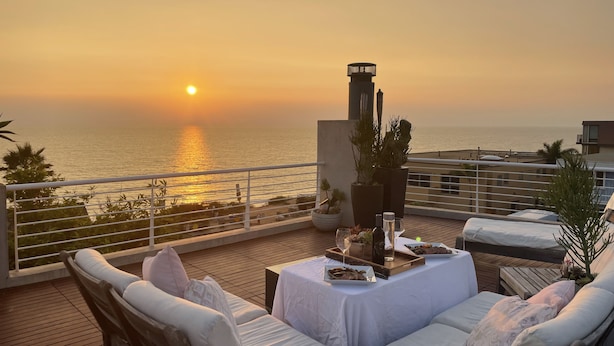 A balcony with a view of the sunset over the ocean, furnished with white cushioned seating and a small table with a bottle and glasses. - Modern Del Mar Beach Home (Del Mar)