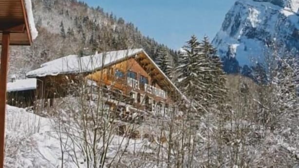 Eine Holzhütte mit verschneitem Dach, umgeben von Bäumen und einem Berg im Hintergrund.