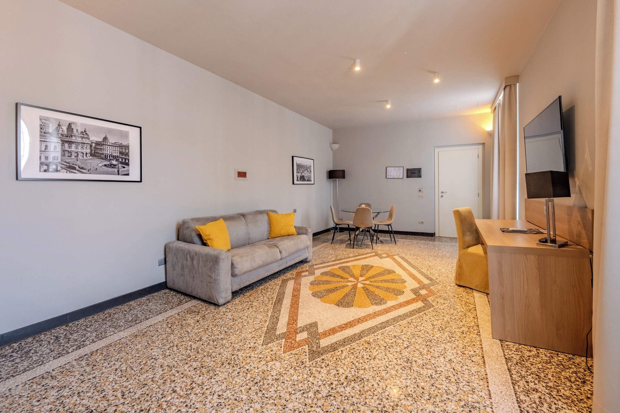 Una sala de estar moderna con un sofá, una mesa de comedor y sillas. Hay un televisor sobre un mueble bajo, una alfombra con un diseño geométrico y cuadros en la pared.