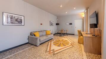 Una sala de estar moderna con un sofá, una mesa de comedor y sillas. Hay un televisor sobre un mueble bajo, una alfombra con un diseño geométrico y cuadros en la pared.