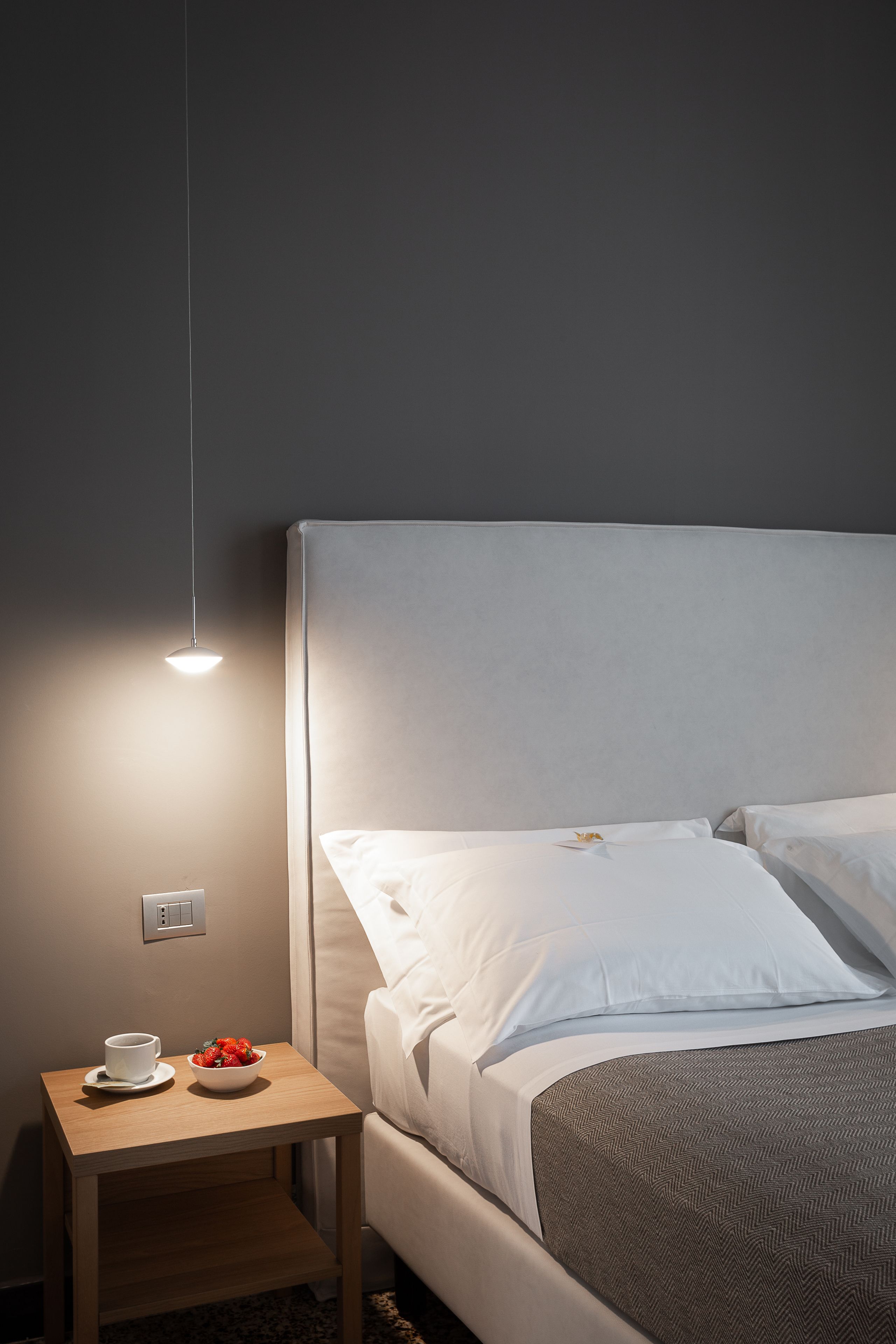 A neatly made bed with white linens and a grey patterned blanket, a bedside table with a cup and bowl of fruit, and a hanging light fixture.