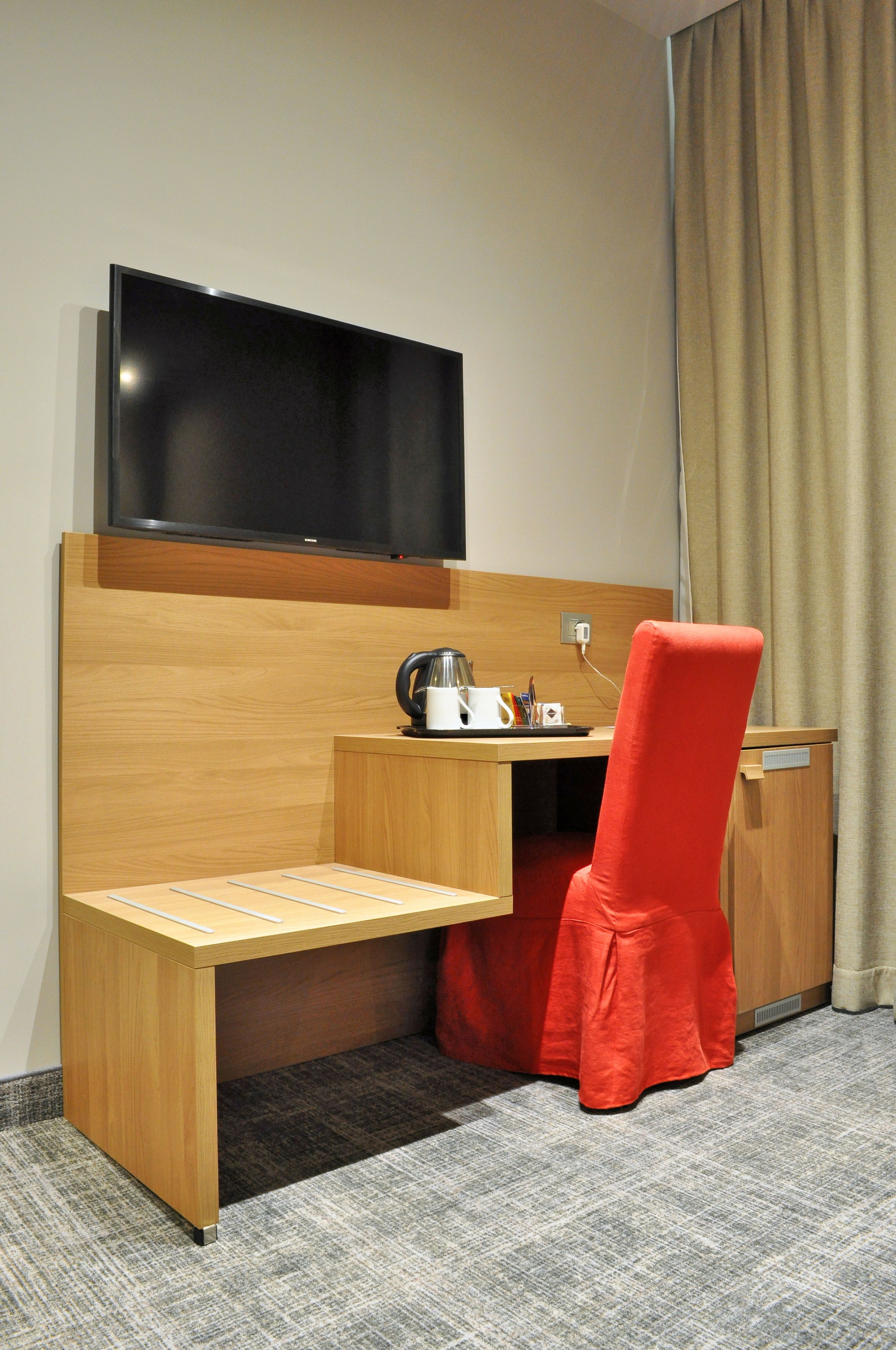 Une chambre d’hôtel avec un bureau en bois, une chaise rouge, un téléviseur à écran plat et une petite table sur laquelle se trouvent une bouilloire et des tasses.