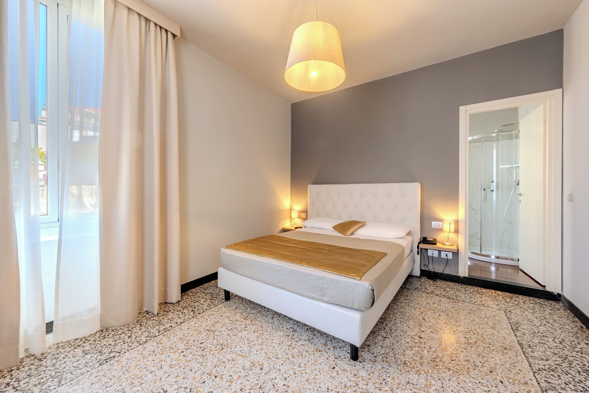 Une chambre d’hôtel avec un lit, des lampes de chevet, une douche en verre et une fenêtre avec des rideaux donnant sur l’extérieur.