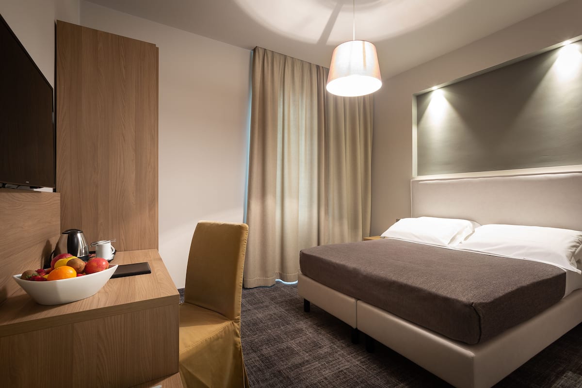 Une chambre d’hôtel avec un lit, un bureau sur lequel se trouve un panier de fruits, une chaise, une machine à café et une grande fenêtre avec des rideaux.
