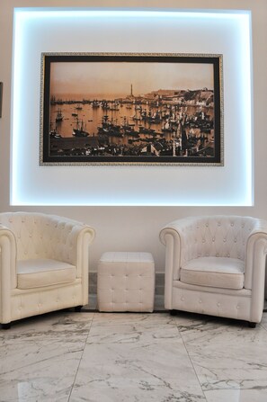 A framed black and white photograph of a harbor with boats, displayed above two white tufted chairs and a matching ottoman on a marble floor. - HNN Luxury Suites (Genoa)