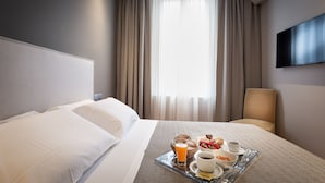 Una habitación de hotel con una cama, una bandeja de desayuno con croissants, fruta, café y jugo, un televisor y un equipo de aire acondicionado.