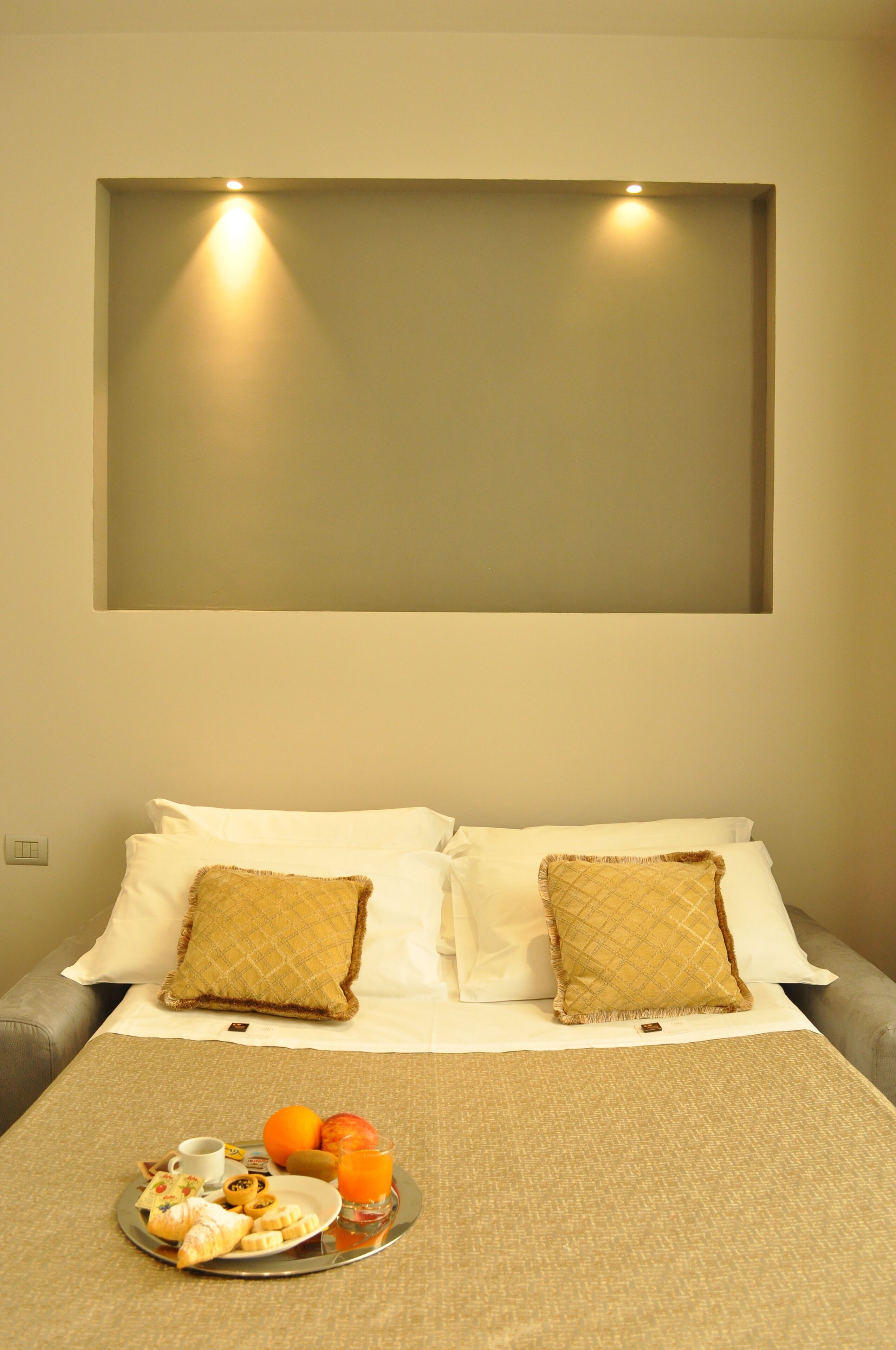 A neatly made bed with two pillows, a tray of breakfast including croissants, fruit, and orange juice, and a wall-mounted light fixture.