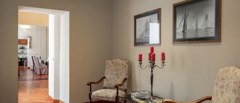 Una habitación amplia con suelo de madera, dos sillas ornamentales, una mesa con candelabro y obras de arte enmarcadas en las paredes.