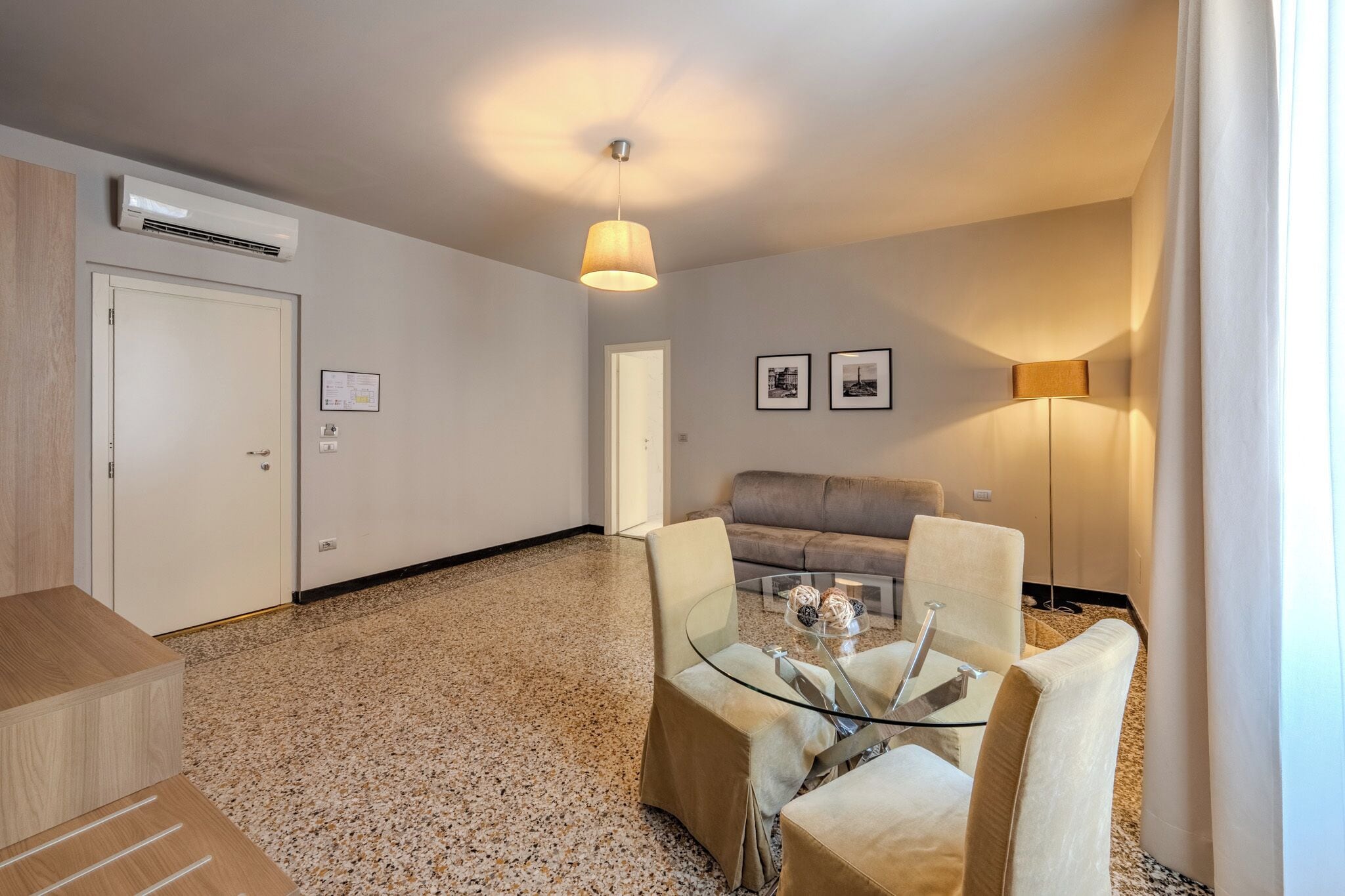 Une chambre d’hôtel équipée d’une kitchenette, d’un canapé, d’une table à manger en verre avec des chaises et d’un luminaire au plafond.