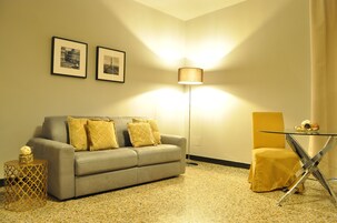 Un salón con un sofá gris, cojines amarillos, una mesa de centro de vidrio, una lámpara de pie y dos cuadros en la pared.