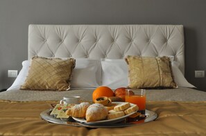 A neatly made bed with a tufted headboard, two pillows, a tray with croissants, a plate with fruit, a glass of orange juice, and a cup of coffee. - HNN Luxury Suites (Genoa)