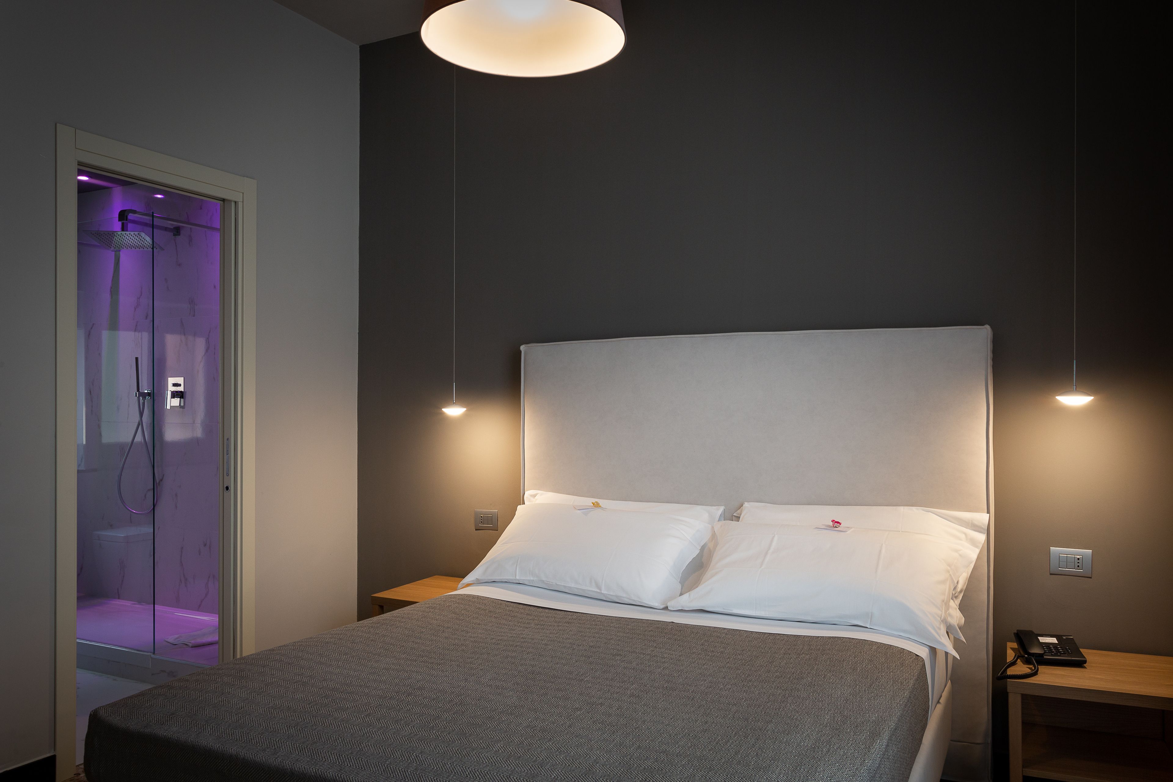 A neatly made bed with white linens, a bedside table with a phone, and a shower area with purple lighting.