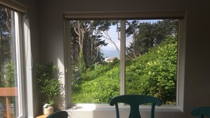 A room with a large window offering a view of a hillside with trees and a distant view of the ocean. There is a wooden table with two chairs and a potted plant on the windowsill. - 3 Bedroom Haven By The Sea w/ beautiful ocean views & 2 Masters Sleeps 10 (Lincoln City)