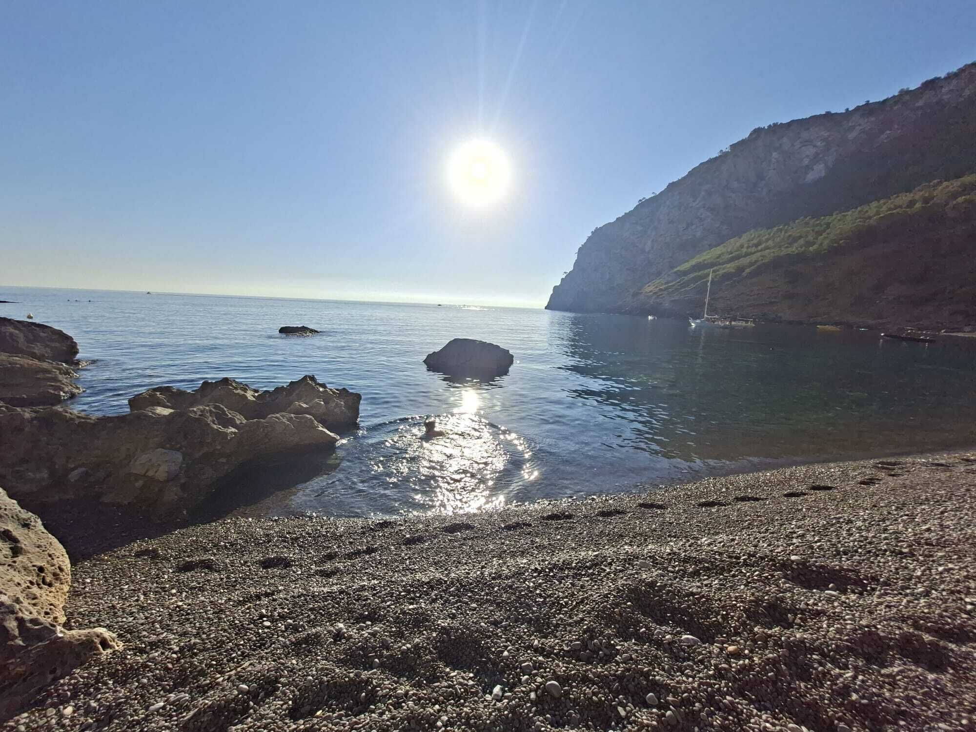 A rocky shoreline with calm water, a clear sky, and a distant sun.