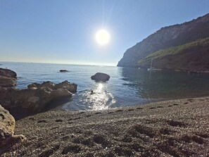Uma costa rochosa com águas calmas, céu claro e um sol distante.